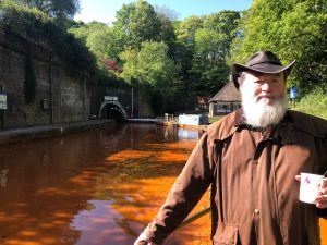 Ein Mann in einem geölten Mantel mit Lederhut am Tiller eines Narrowboots am Ausgang des Harecastle Tunnel in England