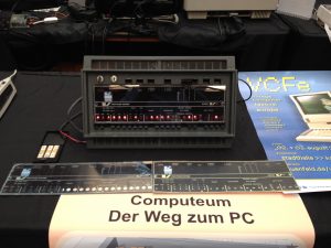 Ein Tisch mit schwarzem Tischtuch, darauf das Four Phase System 70 Forntpanel, davor ein Plakat mit einer Übersicht des Ausstellungsraums im Computeum