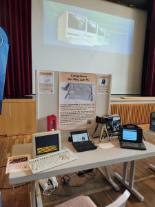Stand mit einem kleinen Linux Notbook, einem Loewe MultiTel und einem VT320 auf einem Tisch, dahinter eine Stellwand mit einem Plakat das das Computeum zeigt.