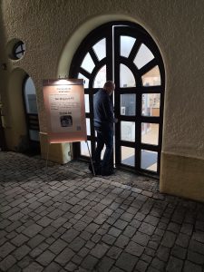 Nächtliches Foto. Das Computeum ist hell erleuchtet. Das licht strahlt durch die Fenster der Türflügle auf die Straße. Vor der Türe steht ein Ständer mit der Einladung zum Besuch, beleuchtet von einem eigenen Strahler. Die Tür zum Computeum wird für Besucher geöffnet. Josef öffnet gerade die Türe für Besucher.