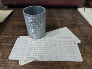 Eine Druckform für Lochkarten steht auf einem Holztisch zusammen mit zwei Lochkarten, einer Standardkarte und einer mit Beschriftung für eine Anwendung. Die Druckform ist ein kleiner Holzylinder aus Aluminium der auf der Aussenseite das Druckbild erhaben trägt.
