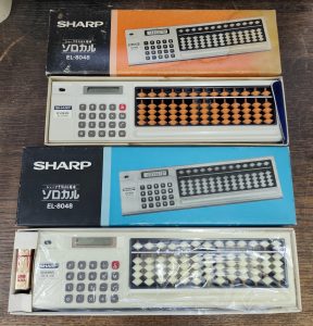 Auf einem Holztisch liegen zwei geöffnete Schachteln. Eine mit schwarz/blauem Design und eine mit schwarz/orangenem. Die Blaue ist etwas länger. In jeder liegt ein EL-8048, in der Blauen zusätzlich eine 9 Volt Batterie. Der El-8048 ist ein weißes, stabförmiges Gerät, ähnlich einem dicken Lineal. Er ist ca 40 cm lang und 5 cm tief. Im linken Viertel ist einfacher 4-spezien Rechner mit Speicher und LCD Anzeige. Der Restliche Raum wird von einem Abakus mit 13 Stegen mit je 4+1 Kunststoffperlen eingenommen. Die Perlen des Rechners in der blauen Schachtel sind Weiß, die des Abakus in der orangen Schachtel sind Holzfarben. In der Orangen Schachtel sieht man noch einen Zipfel einer beiliegenden Schutztasche aus blauem, samtenen Stoff.