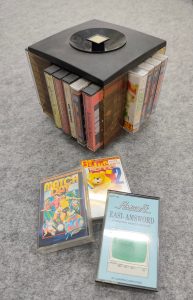 Auf einem grauen Filz steht ein schwarzer würfelförmiger Kasten in dessen Fächer an den vier Außenseiten jeweils fünf Kompaktkassetten hochkant gesteckt werden können. Der Würfel steht auf einem (unsichtbaren) drehbaren Fuß und hat einen versenkten Knopf zum Anheben an der Oberseite. In den Fächern stecken verschiedene Kassetten mit Software, hauptsächlich Spiele. Vor dem Würfel liegen 3 Kassetten. "Match Day", ein Fußball Spiel; "School 2", eine Lernkassette für unter 6-jährige und "Easi-Amsword", eine minimale Textverarbeitung