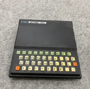 Auf einem grauen Filz liegt ein schwarzer Timex Sinclair 1000. Im Unterschied zum ZX81 fehlt der erhabene Schriftzug ZX81m dafür ist an der oberen Kante ein silberner Aufkleber "Timex Sinclair 1000". Über der Folientastatur ist ein Tastaturaufsatz befestigt der 'richtige' Tastenkappen bietet. Die Tasten sind beige für den Alpha-Teil, hellgrün für die Ziffern und orange für New Line und Shift. Die Beschriftung auf und neben den Tasten ist sauber gedruckt und exakt wie das Original.