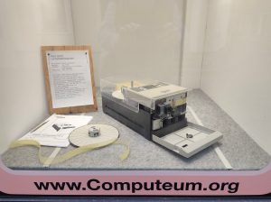 Im Schaufenster des Computeums ist ein Facit 4070 Lochstreifenleser, seine Anleitung und eine Rolle Lochstreifen zu sehen