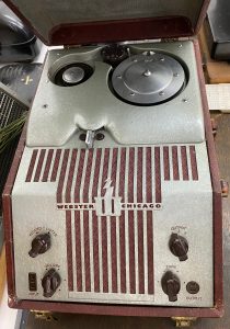 Ein an der Schmalseite geöffneter brauner kofferartiger Kasten mit einem metallenen Einbau. Man sieht hinten eine kleine und eine große Spule. Vorne sind Bedienschalter für Aufzeichnen/Anhören, Lautstärke, Tonlage sowie ein Wahlschalter für 3 mögliche Ausgabewege. Zwischen den Schalten ist ein Gitter sichtbar hinter dem sich ein Lautsprecher befindet. Auf dem Gitter ist "Webster Chicago" sowie ein Logo bestehend aus einem W und einem Blitz zu sehen.