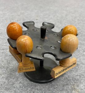 Auf einem grauen Filz steht ein klassisches schwarzes Stempelrondell aus gepresstem Blech. In vier der acht Halterungen hängen Stempel. Auf zweien davon kann man eine Aufschrift sehen: "Skonto" und "Briefdrucksache".