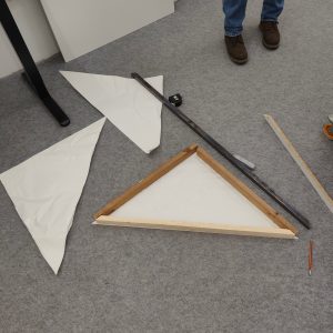 Drei Dreiecke aus Papier auf dem Boden, daneben mehrere Holzlatten Bleistift und Messgeräte