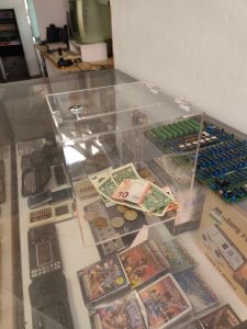 Auf der Vitriene mit den Handheld Computern und Spielgeräten steht eine durchsichtig Plexiglaskiste mit verschiedenen Banknoten und Geldstücken.