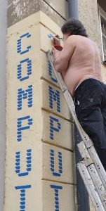 An der Hausecke steht eine hohe Leiter, darauf der Hans der Gerade das C von Computeum an die Wand malt.