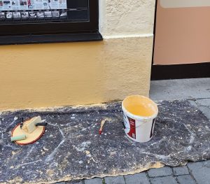 Ein Stück Sims ist zu sehen, davor auf einer Abdeckung aus Malerfilz ein Eimer mit Farbe und Pinsel.