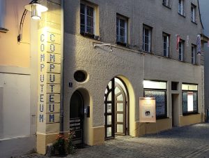 Aussenansicht des Computeums in der Dämmerung. Links an der Ecke ist der neue Schriftzug zu sehen. Im Computeum brennt Licht, die Tür ist auf und ein Schild steht daneben.